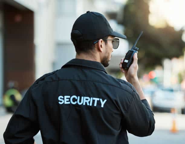 A security guard wearing a black uniform and cap uses a walkie-talkie while standing outdoors. The back of his jacket reads 