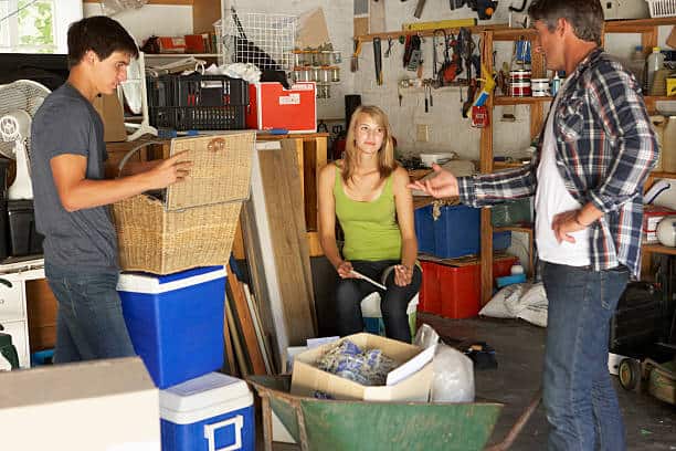 Three people organizing a cluttered garage. A man holds a basket, a woman sits on a stool, and another man gestures with his hand. Various storage items and tools are visible around them.