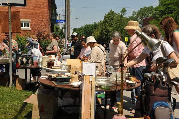 People browsing items at an outdoor yard sale on a sunny day, with tables displaying various goods like lamps, cookware, and golf clubs. Trees and brick buildings are in the background.