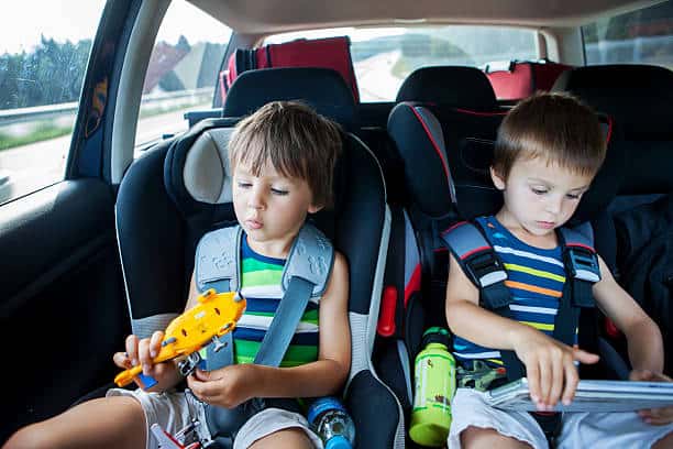 Two young boys sitting in car seats in the back of a vehicle. One is holding a toy, while the other looks at a book. Both are wearing striped shirts.