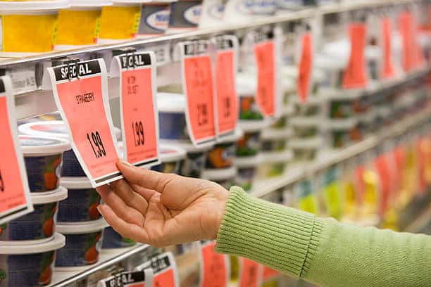 A person in a green sweater reaches for a yogurt container in a grocery store. The shelf is labeled with sale tags displaying prices.