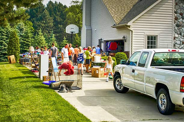 People browsing items at a yard sale on a driveway with a white truck parked nearby.