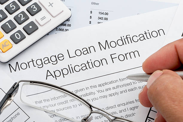 Close-up of a person filling out a Mortgage Loan Modification Application Form; glasses, a pen, and a calculator are visible.