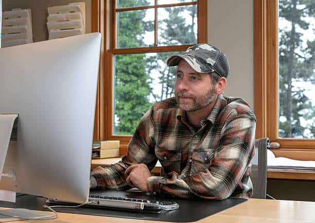 A man in a plaid shirt and cap sits at a desk, looking at a large computer monitor, with windows showing trees in the background.