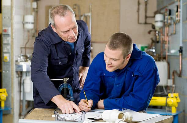 Two men in blue work uniforms focus on documents in an industrial setting. The older man points, while the younger man takes notes. Various pipes and industrial equipment are visible in the background.