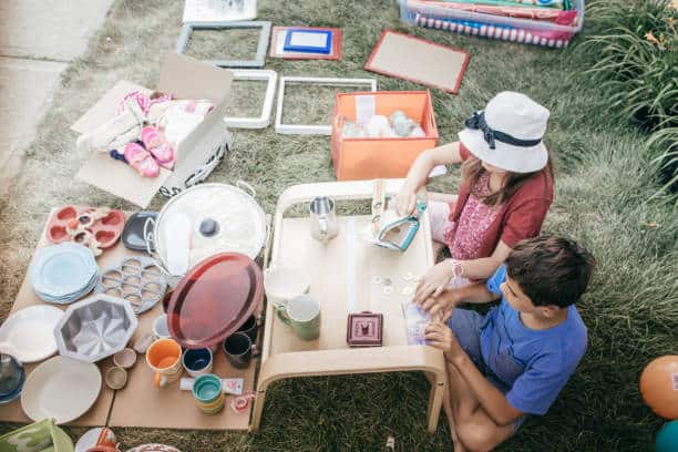 Two children sit at a small table surrounded by various household items and toys on a grassy area. They appear to be organizing or sorting the items.