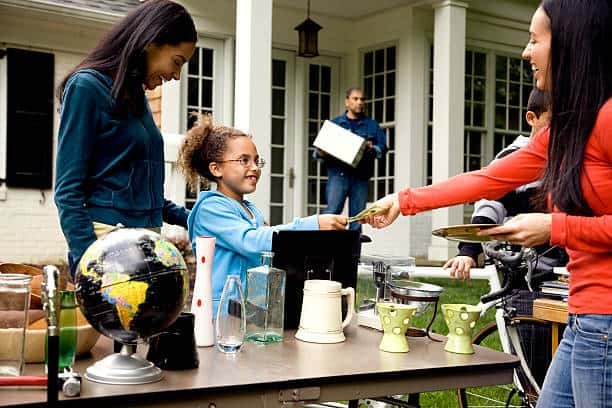 People at a yard sale; a child hands money to an adult across a table with various items, including a globe and dishes.