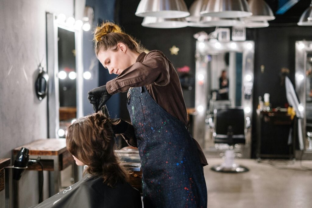 A hairstylist, wearing a dark apron, works on a client's hair in a modern salon with bright lighting and mirrors.