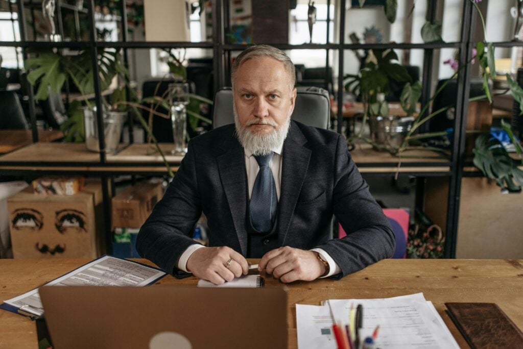 An older man with a gray beard sits at a wooden desk in an office, wearing a dark suit and blue tie, with documents and stationery in front of him.