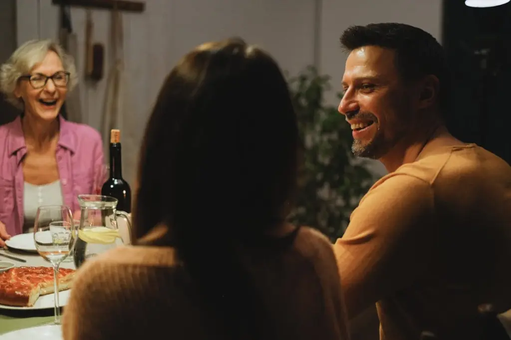 Three people sit at a table, smiling and enjoying a meal together. A bottle of wine and a pizza are visible on the table.