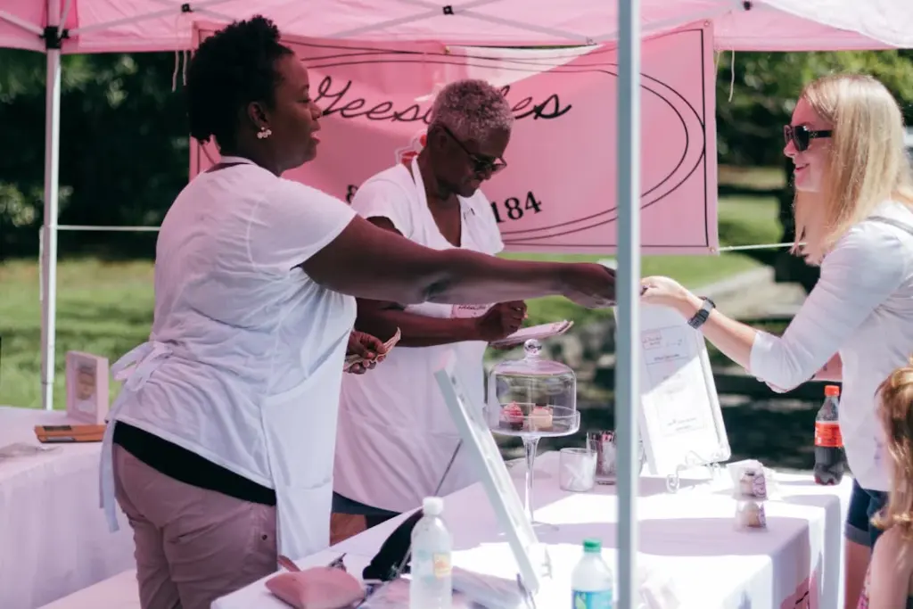 Two vendors in aprons serve a customer at an outdoor market booth with a pink sign and baked goods on display.