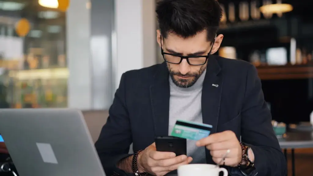 A man sitting at a desk in a cafe is looking at his smartphone while holding a credit card, with a laptop open in front of him.