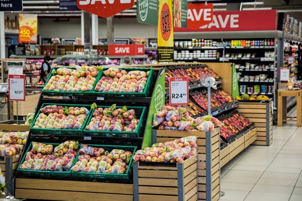 Grocery store interior featuring displays of bagged apples in the foreground, with various packaged goods and fresh produce on shelves and stands in the background. Signs indicate sale prices, catering to the budget-conscious lower middle class community.