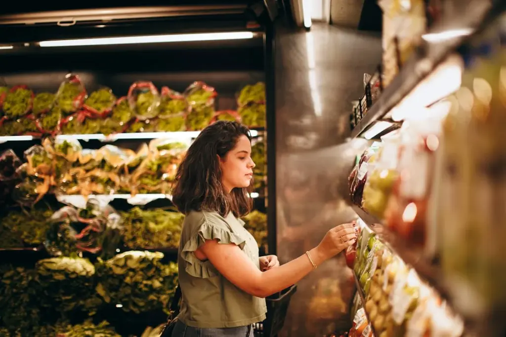 A woman selects produce from a grocery store shelf, surrounded by various leafy greens.