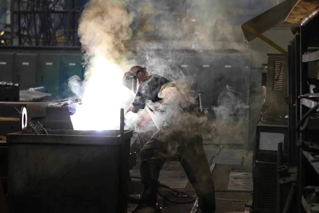 A worker in protective gear is welding with bright light and smoke emitting from the process in a dimly lit industrial workshop.