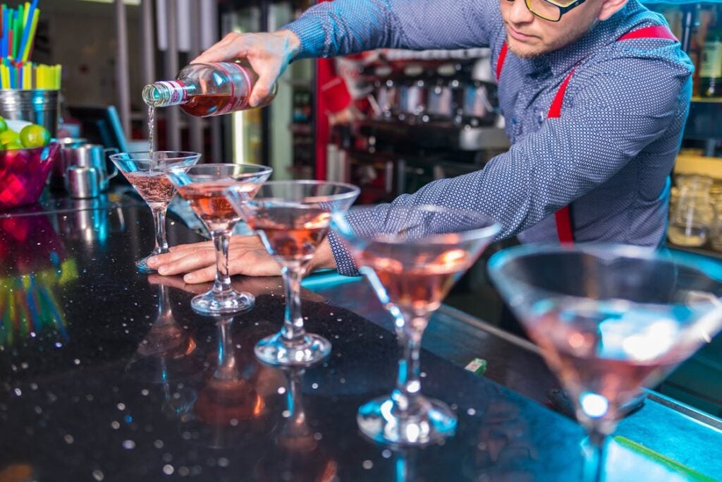 A bartender wearing glasses and a blue shirt with red suspenders is pouring a pink liquid into martini glasses lined up on a bar counter.