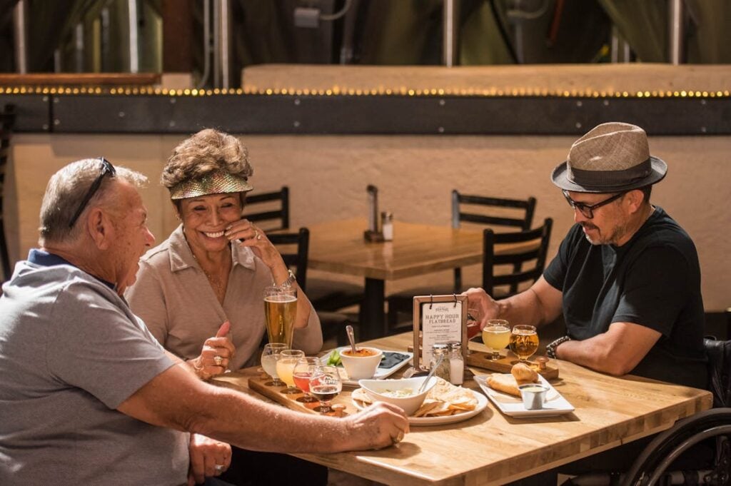 Three people are seated at a restaurant table, talking and enjoying food and drinks. Two men and a woman with a drink in hand are engaged in conversation, surrounded by chairs and a menu on the table.