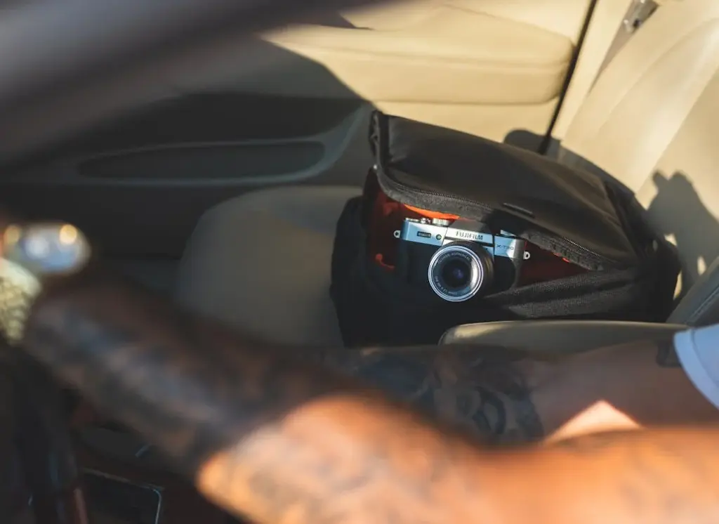 A camera partially visible in an open backpack on a car seat, with a tattooed arm in the foreground.