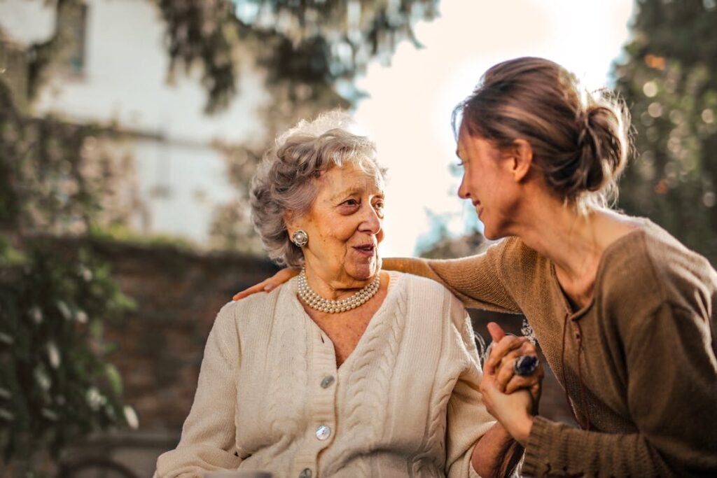 An elderly woman with gray hair and a young woman with brown hair sit close together outdoors, smiling and holding hands, cherishing the moment away from the bad effects of digital technology.