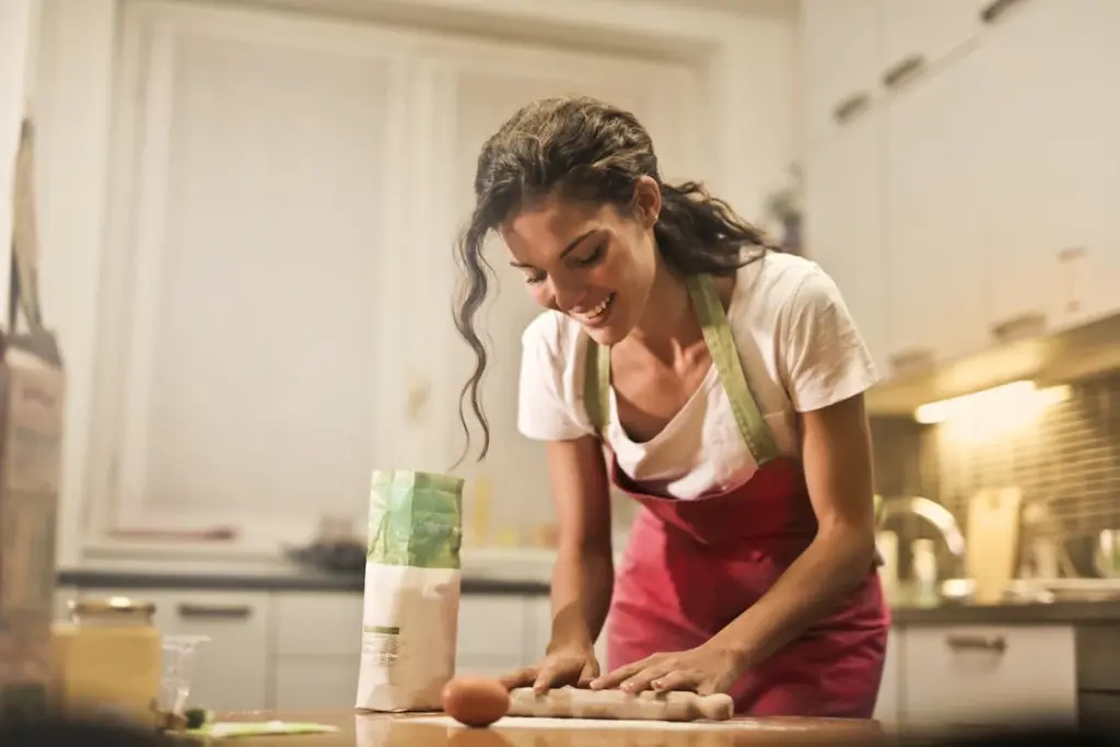 A woman in a pink apron smiles while rolling out dough in a kitchen, with an egg and a bag of flour on the counter.