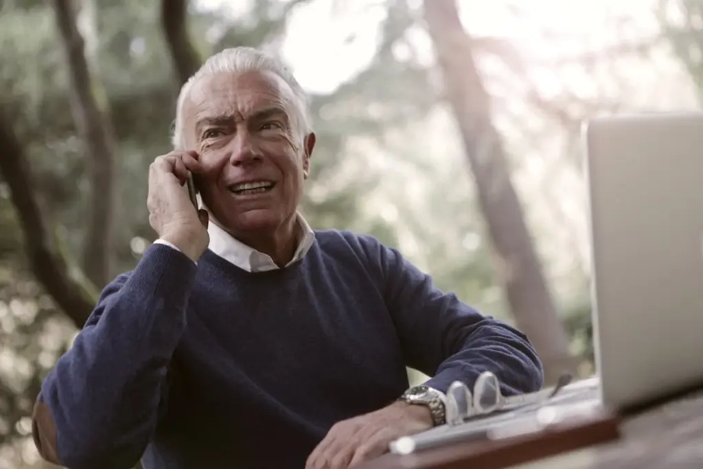 Older man in a blue sweater talks on a smartphone while sitting outdoors at a table with a laptop and eyeglasses.