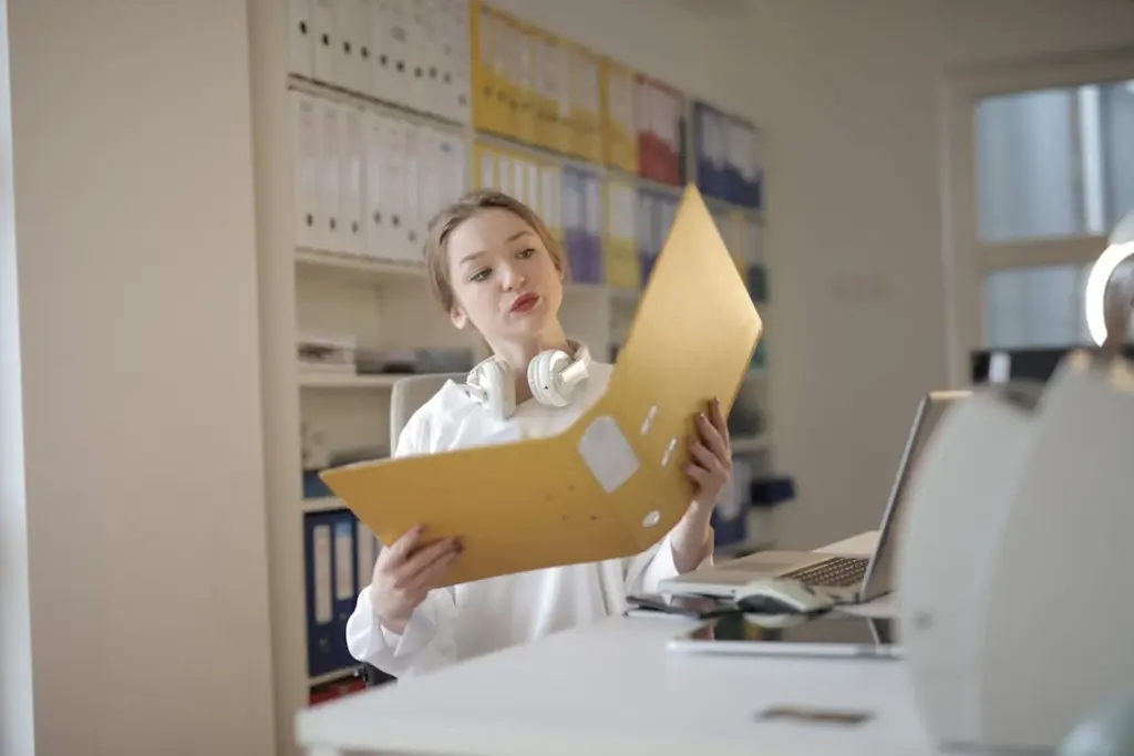 A woman in a white shirt reviews a large folder while sitting at a desk in an office environment, surrounded by filing cabinets and office supplies.