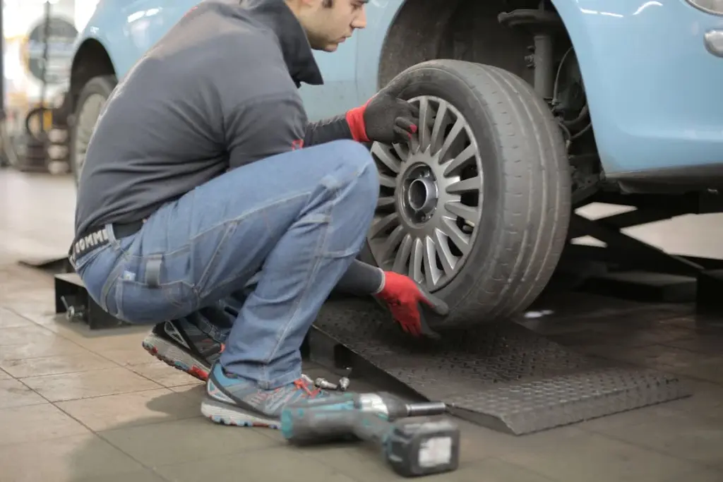 A mechanic in jeans and red gloves replaces a car tire on a raised platform using a power tool.