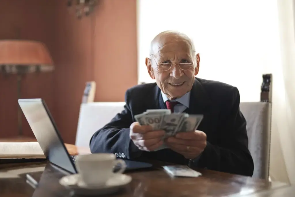 An elderly man in a suit sits at a table with a laptop, looking at a stack of dollar bills with a smile, reflecting on how life priorities shift with age. A cup and a lamp are visible in the background.