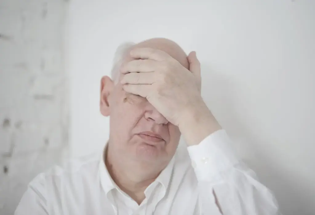 An elderly man wearing a white shirt covers his face with his hand, showing an expression of frustration or stress against a plain white background.