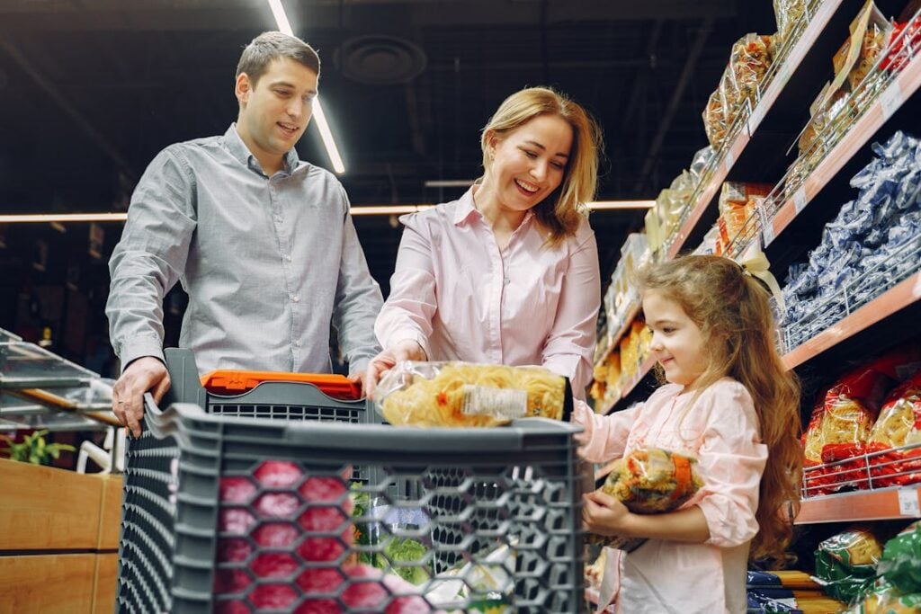 A man, woman, and girl are shopping in a grocery store. The girl is holding a package of food and placing it into the shopping cart. Shelves filled with products are visible in the background.