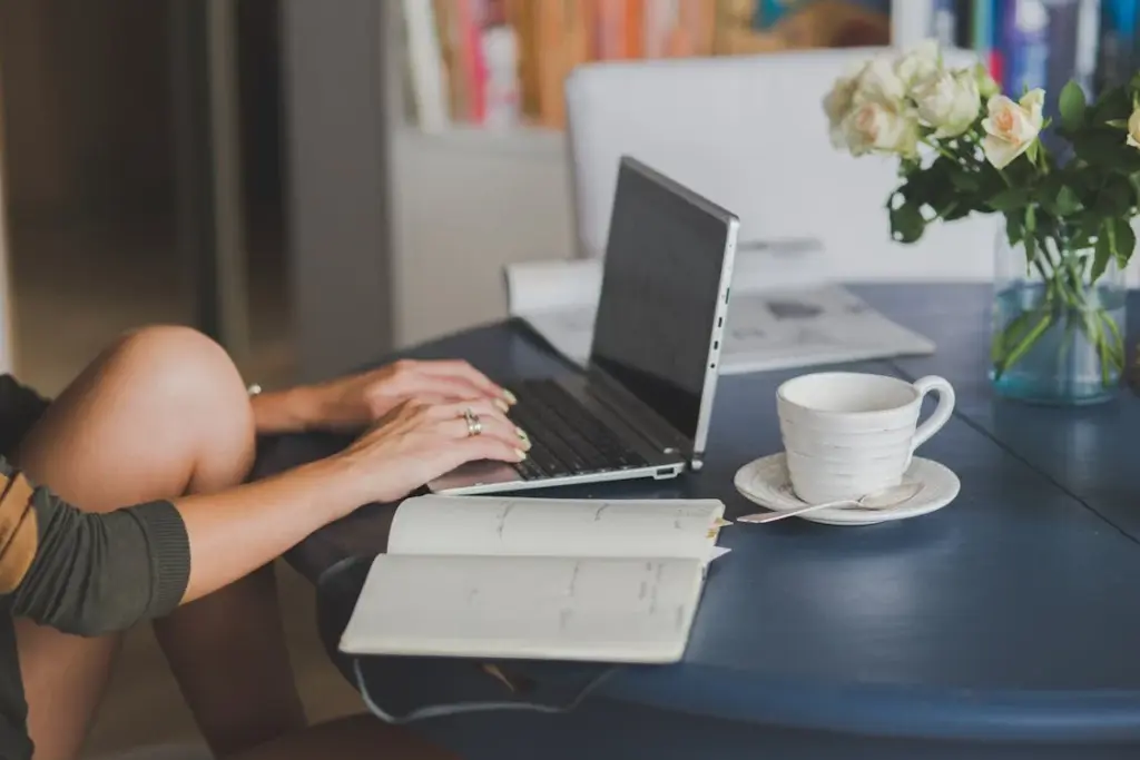 Person typing on a laptop at a table with a notebook, cup of coffee, and a vase of white roses.