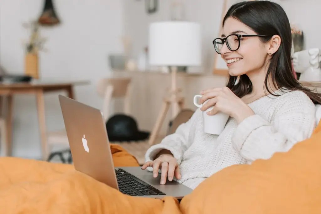 A woman with glasses sits on a couch, holding a mug, and uses a laptop. She is covered with a yellow blanket, and there is a lamp and a chair in the background.