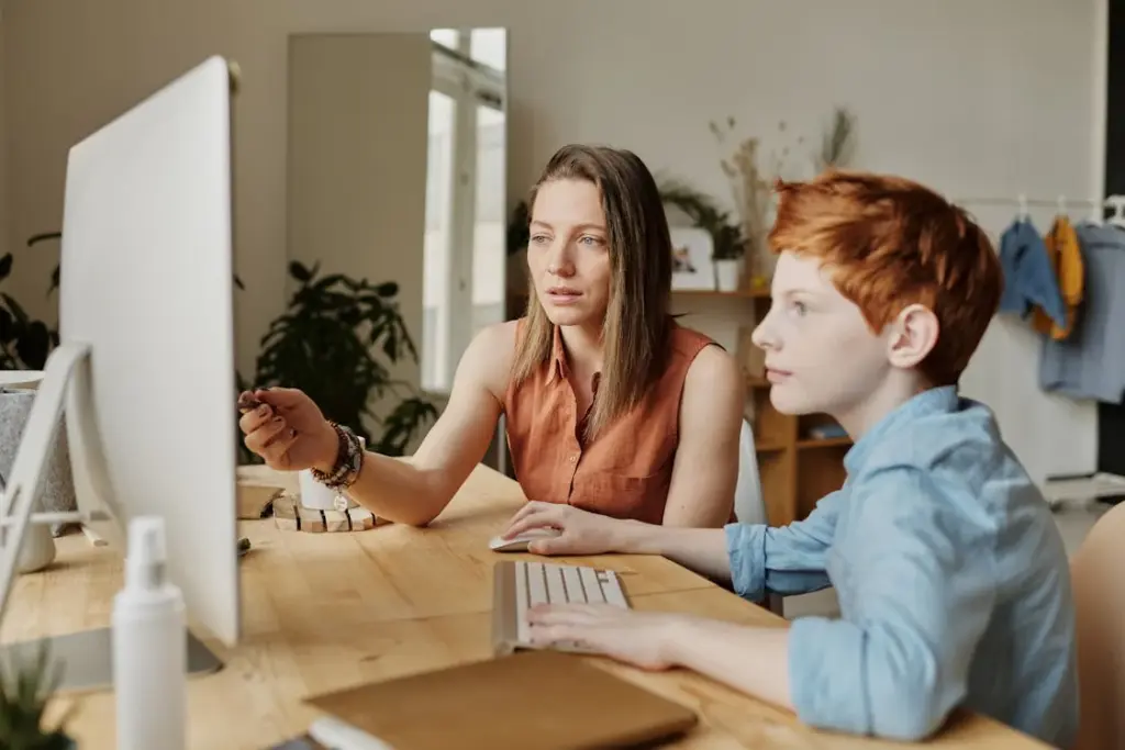 A woman and child sit at a wooden desk using a computer together in a bright room.