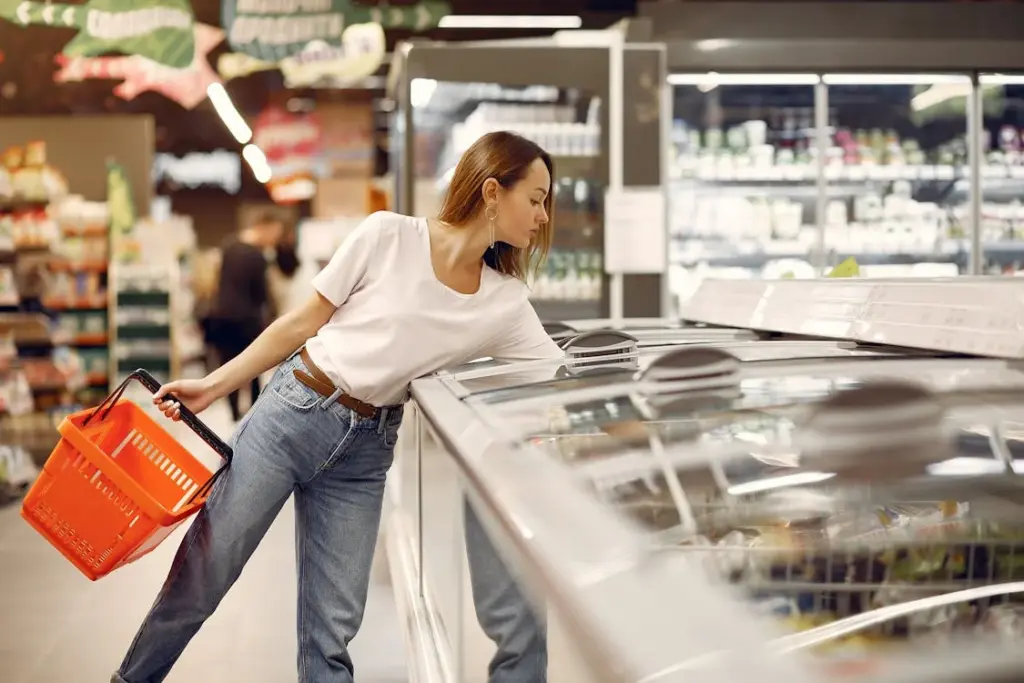 Person holding a red shopping basket while reaching into a freezer in a grocery store.