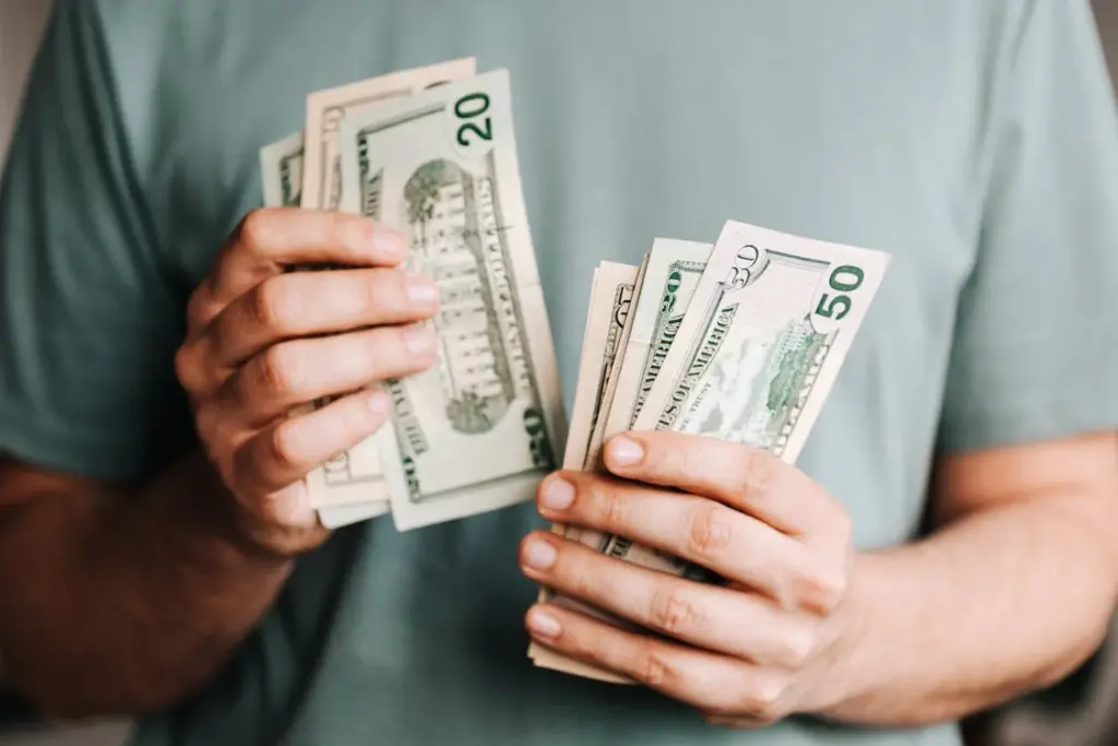 A person in a grey shirt is holding multiple U.S. dollar bills in both hands.