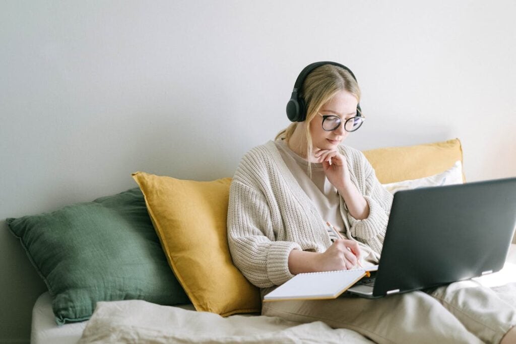 A person wearing headphones and glasses is sitting on a bed, writing in a notebook while looking at a laptop. They are surrounded by pillows.