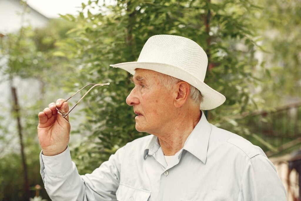 An elderly man wearing a white hat holds a pair of glasses while looking to the side, with greenery in the background.