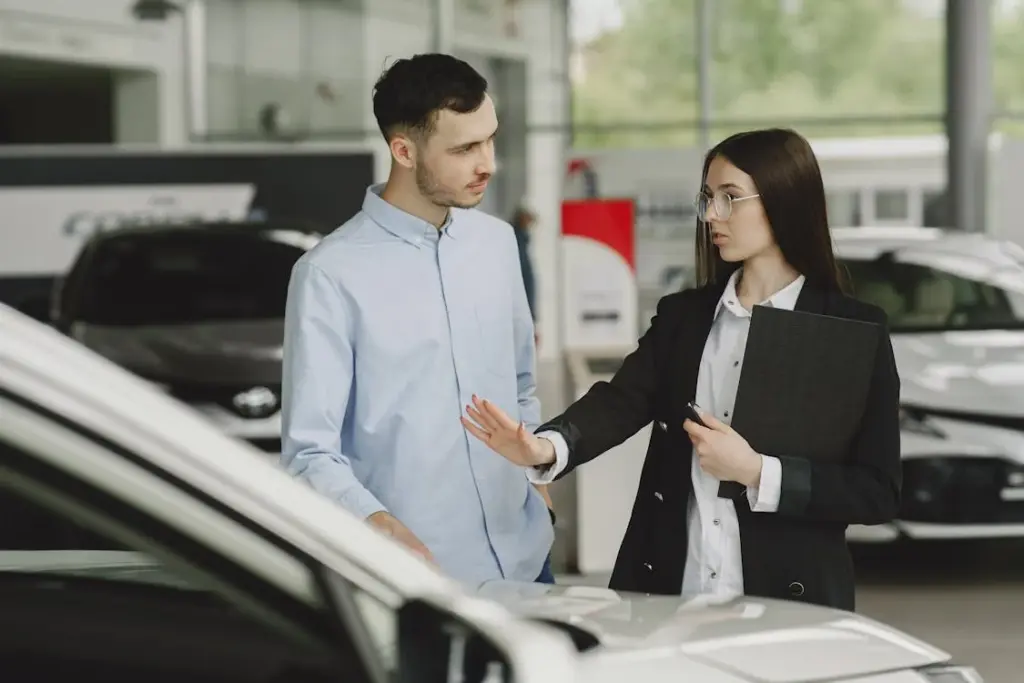 A car salesperson, holding a clipboard, talks to a potential buyer in a showroom as they stand next to a white car.