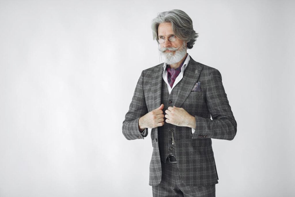 Elderly man with gray hair and beard in a plaid three-piece suit adjusting his jacket, standing against a plain white background, illustrating the misguided reasons why companies do not hire over 50.