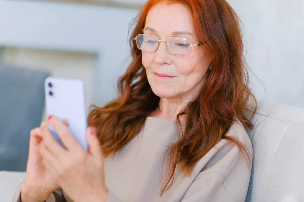 Elderly woman with glasses and red hair sits on a couch, using a smartphone.