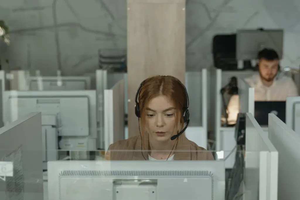 A woman wearing a headset works on a computer at a cubicle in an office. Another person is visible working in the background.