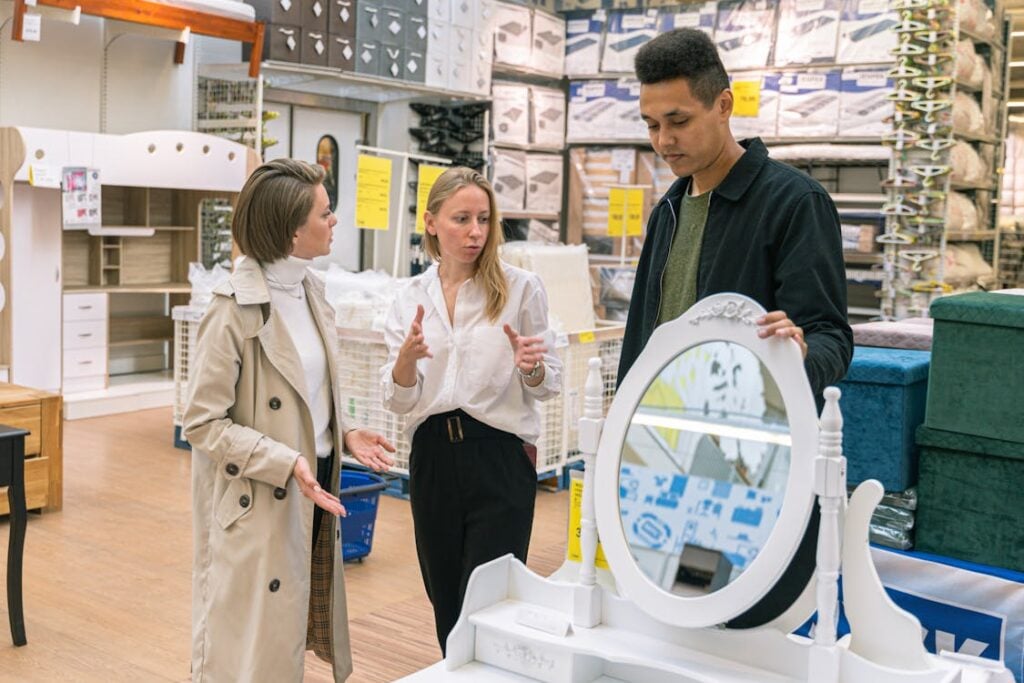 Three people are discussing in a store with furniture and beddings in the background. One person gestures while speaking, the other two listen, standing next to a white vanity table with a round mirror.