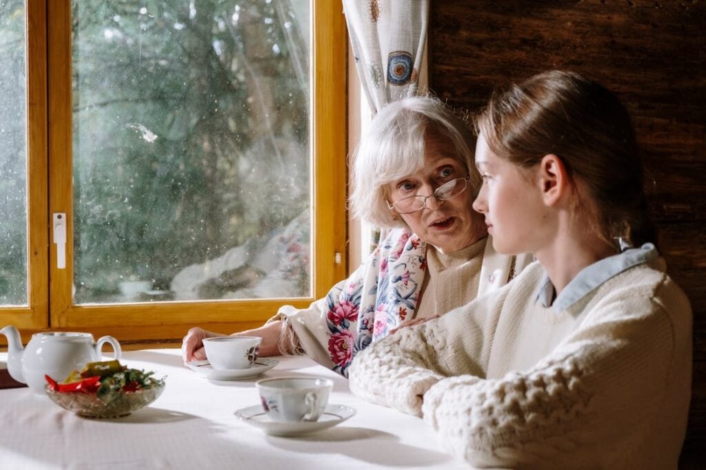 An elderly woman and a younger woman sit at a table with teacups and a teapot, talking by a window with a curtain.