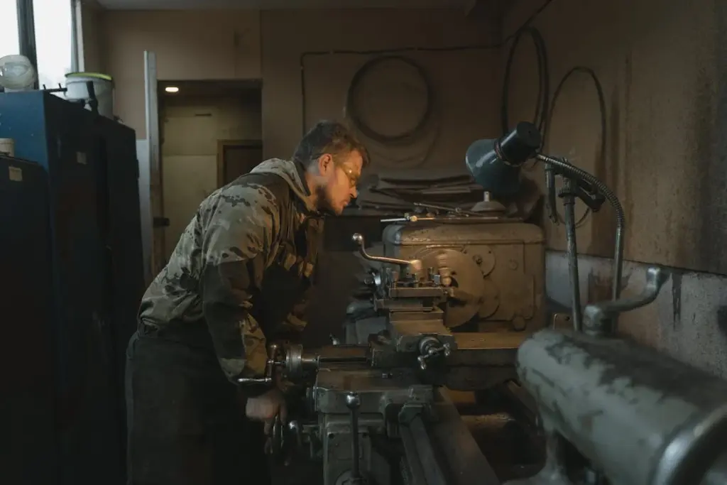 A man operates a lathe machine in a dimly lit workshop, wearing protective gear and focusing intently on his work.