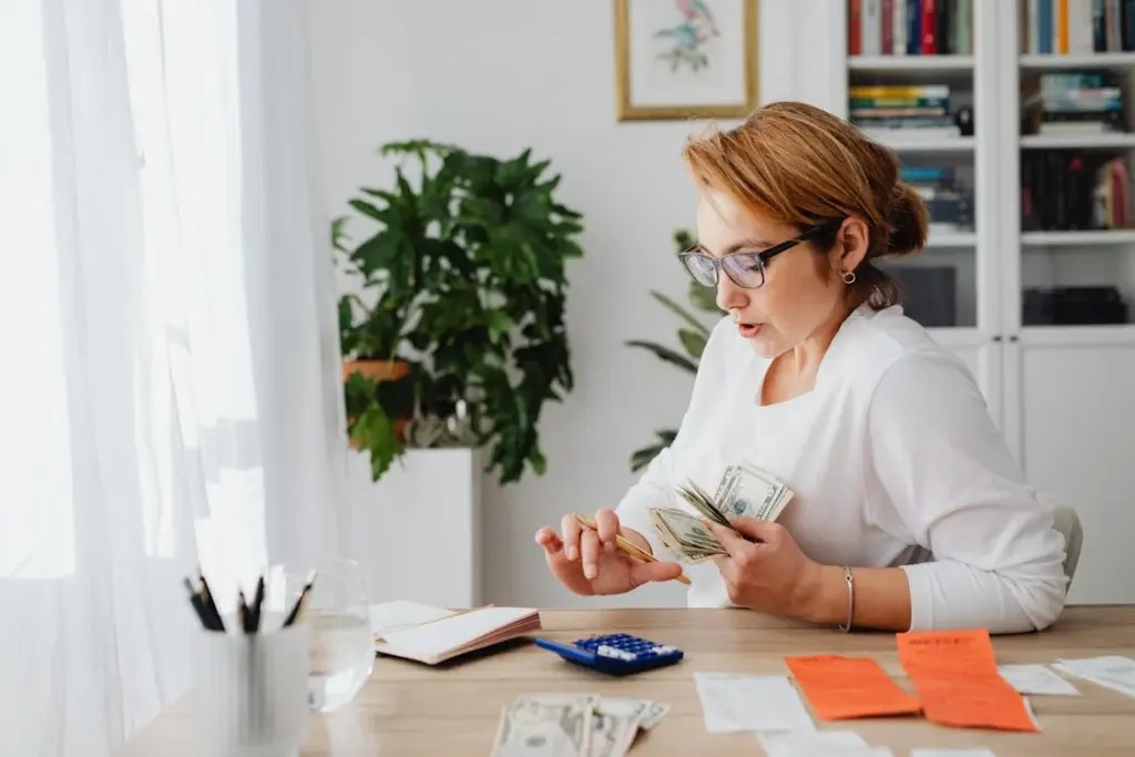 A woman counts money at a desk, with a notebook, calculator, receipts, and a glass of water nearby.