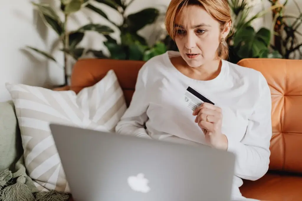 A woman in a white shirt sits on a couch with plants in the background, holding a credit card and looking at a laptop.