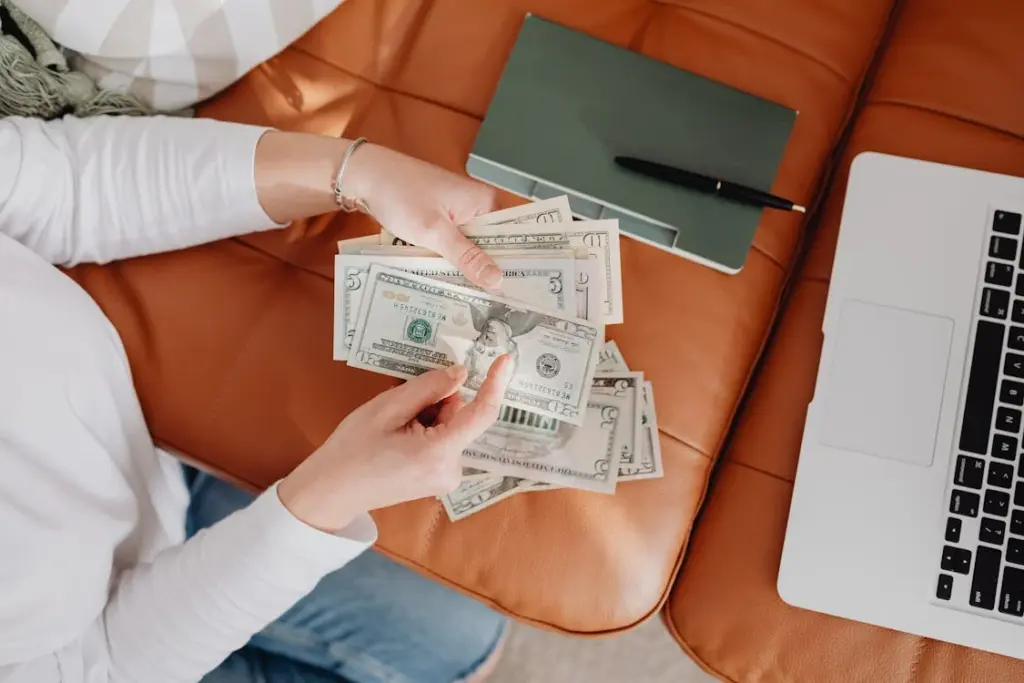 A person counts dollar bills while sitting on a brown couch next to an open laptop and a closed green notebook with a pen.