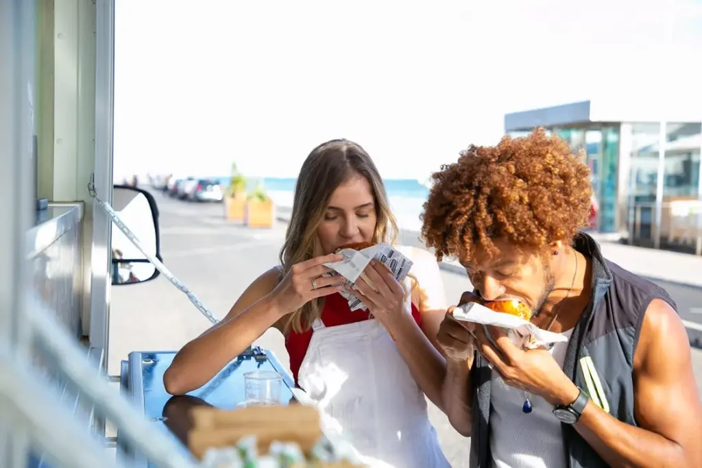 Two people eating sandwiches near a food truck at an outdoor location.