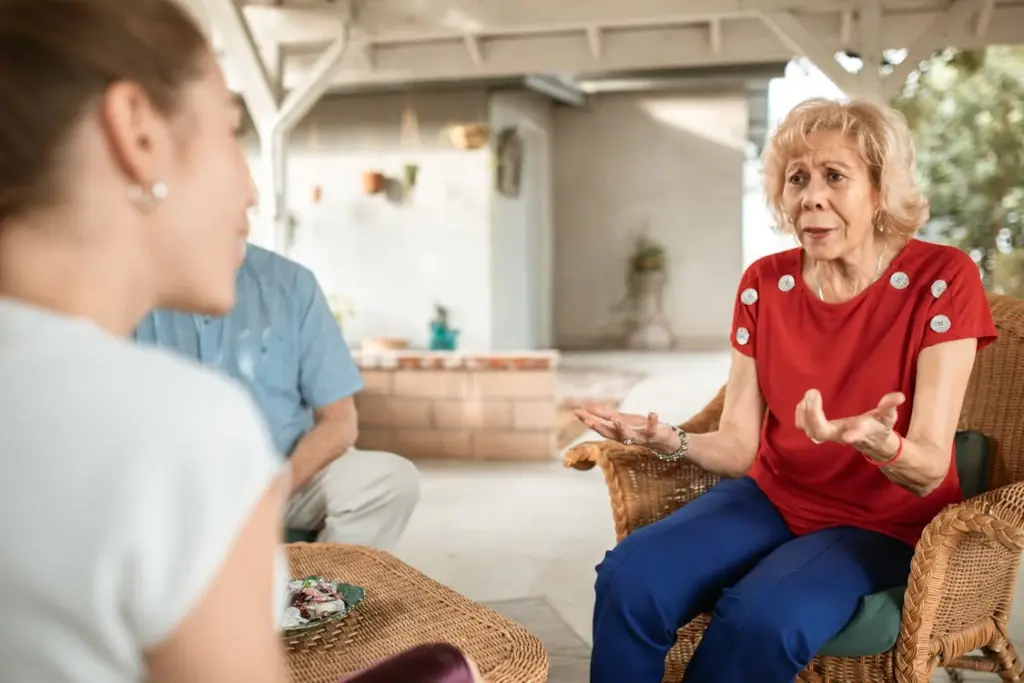 An elderly woman in a red shirt gestures with her hands while talking to a seated younger woman. They are outdoors under a covered structure.