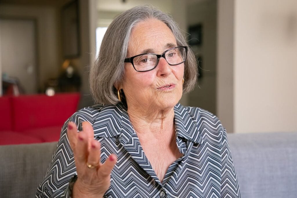 An elderly woman with short gray hair and glasses sits on a couch, gesturing with one hand. She is wearing a patterned blouse and appears to be speaking.